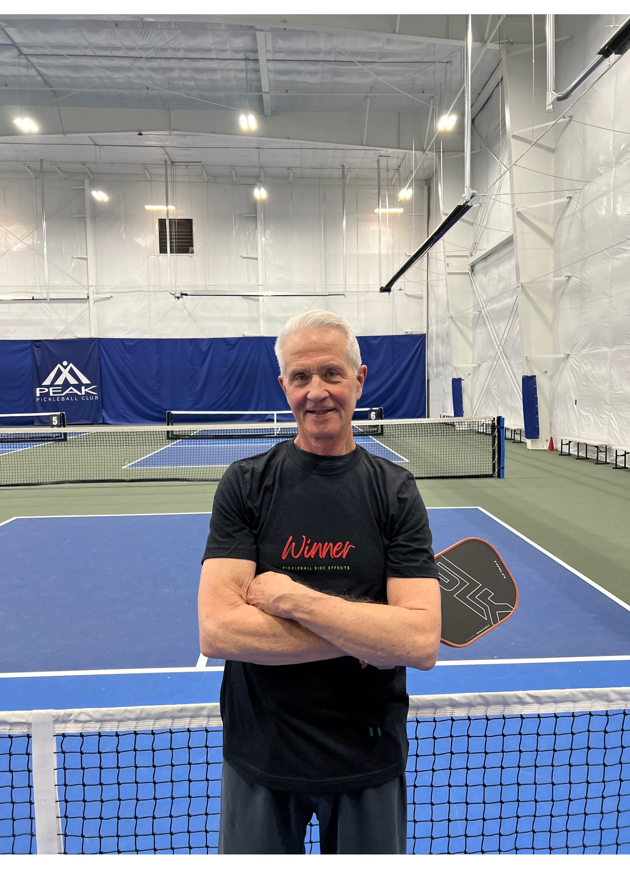 George wears a Winner, Pickelball Side Effects T-shirt.  Winner is in Red letters and a nice script writing and the other words are green.