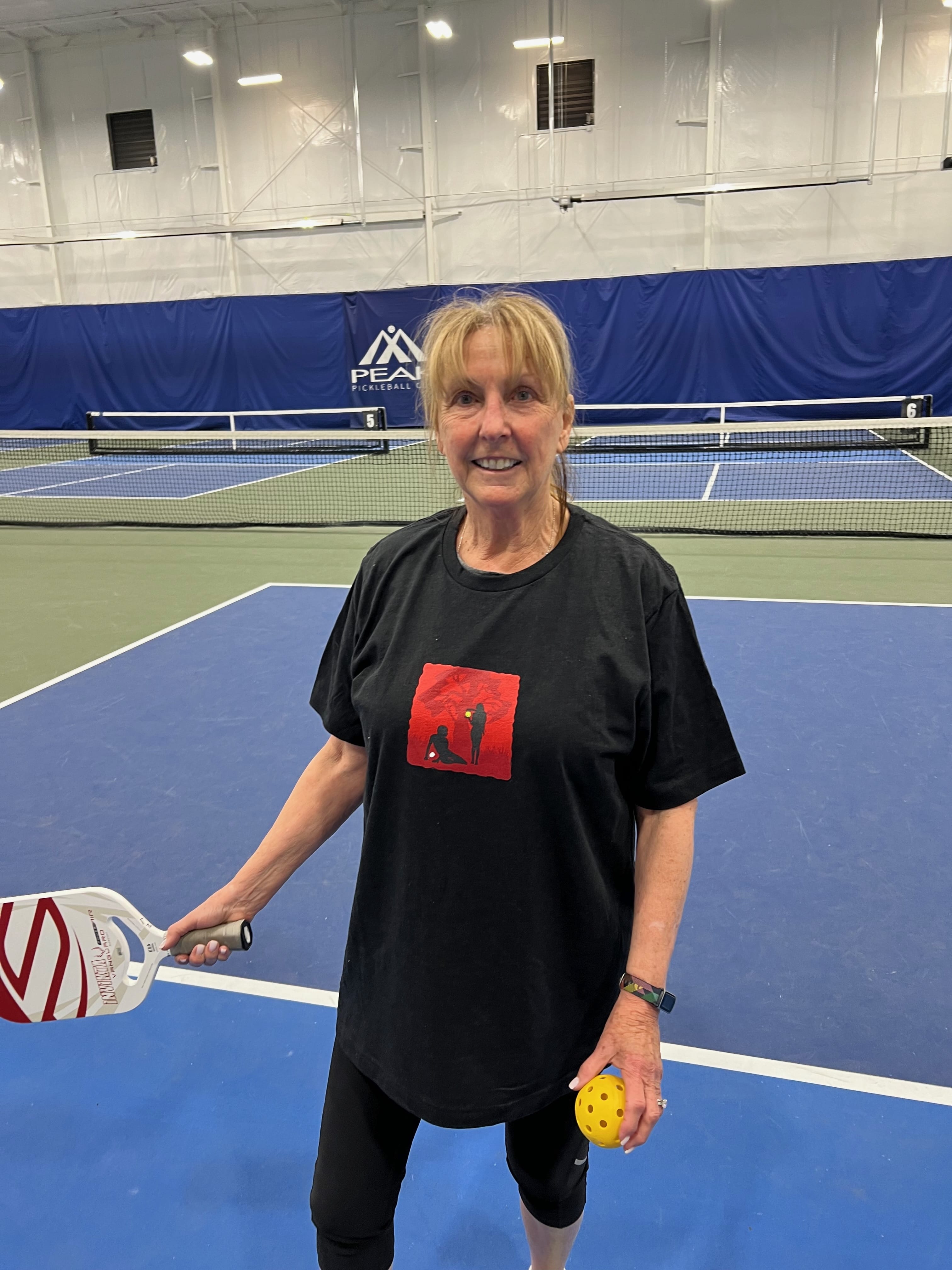 Lynda poses in a Unisex Pickleball t-shirt with an image only of Adam and Eve with a pickleball and a paddle